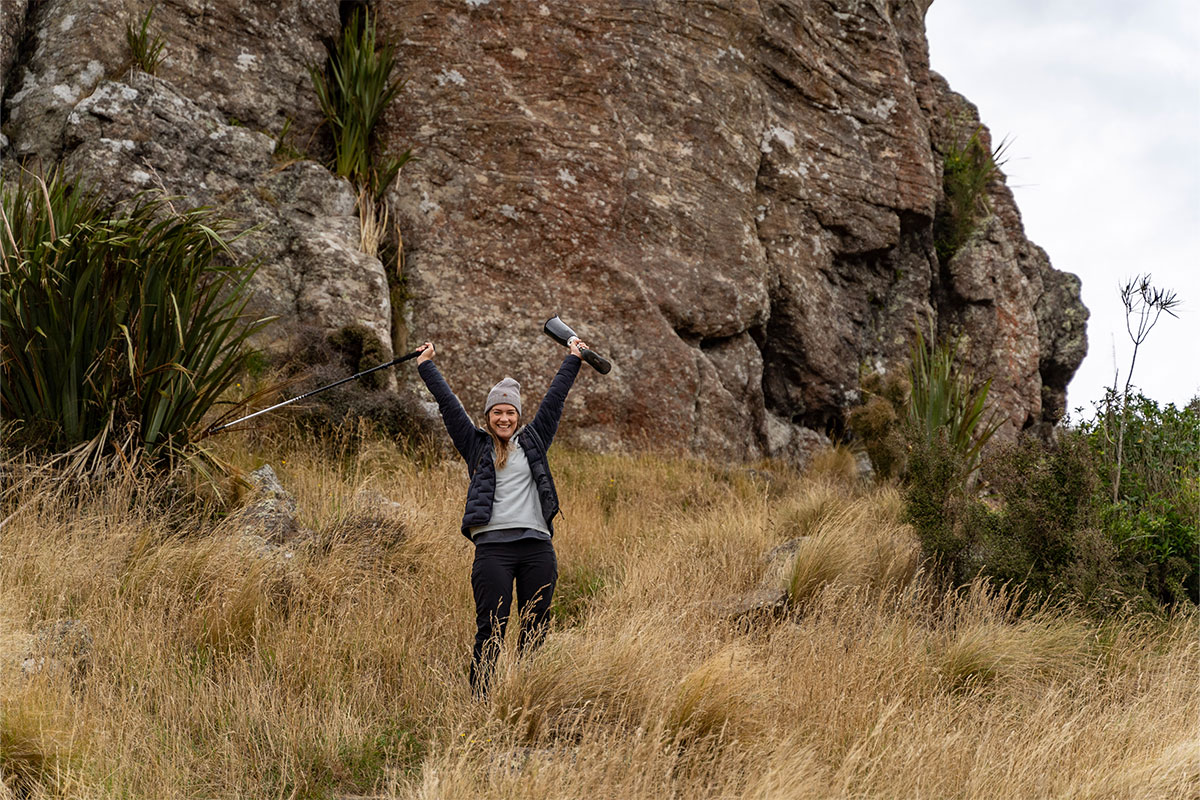 Rachel Maia smiling with her prosthetic leg in the air against the backdrop of Lyttelton Crag, Christchurch, New Zealand